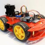 A close-up view of a four-wheeled robot car setup. The chassis is red, with yellow wheels. An Arduino board, an L298N motor driver, an HM-10 Bluetooth module, and wiring are mounted on the chassis. This setup appears to be designed for remote-controlled robotic movement, likely controlled via a Bluetooth app.