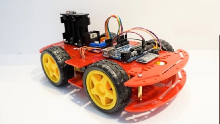 A close-up view of a four-wheeled robot car setup. The chassis is red, with yellow wheels. An Arduino board, an L298N motor driver, an HM-10 Bluetooth module, and wiring are mounted on the chassis. This setup appears to be designed for remote-controlled robotic movement, likely controlled via a Bluetooth app.